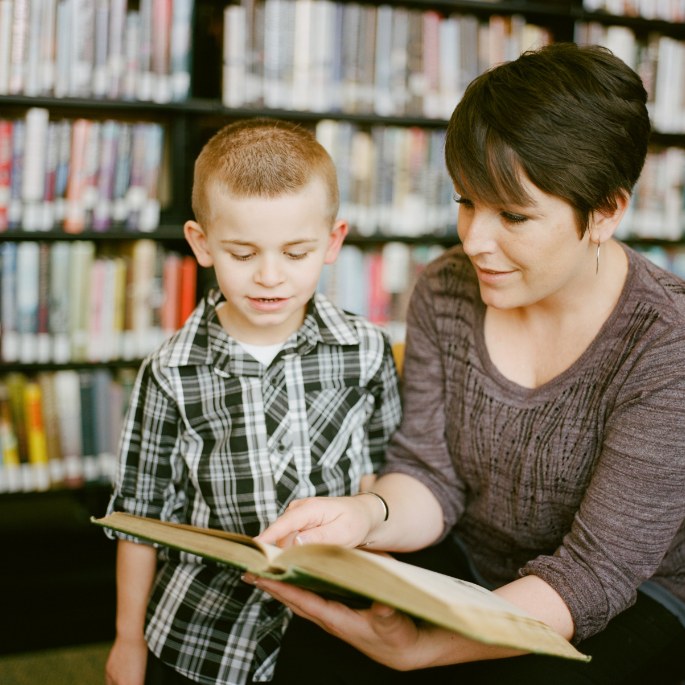 A woman sitting on a stool in the library reading a book. There is a young boy standing beside her looking at the book.