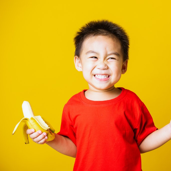 A smiling young boy in a red t-shirt holding a half peeled banana.