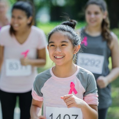 A child, a teen and a woman all running with a numbered bib on them. They all have bright pink breast cancer support ribbons pinned on their tops.