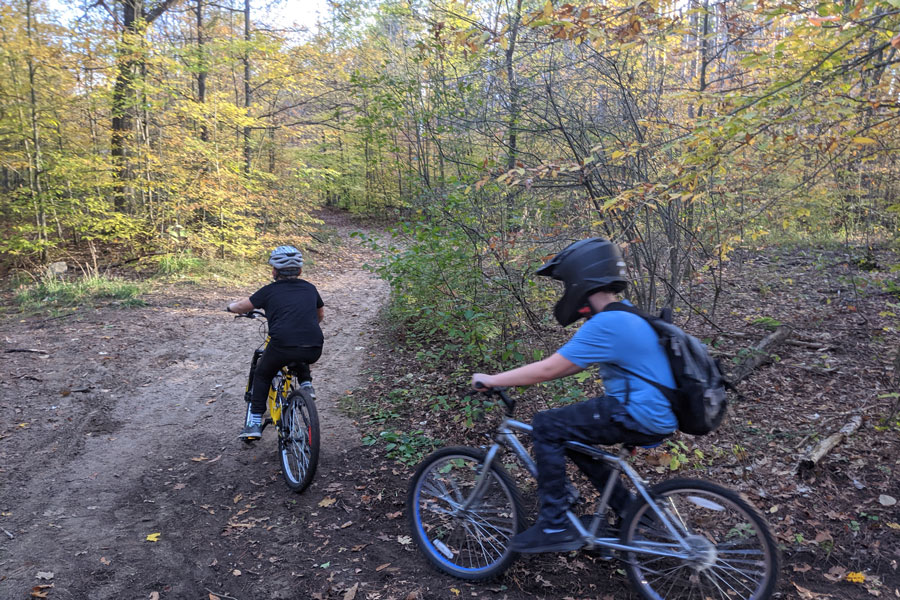 Two students riding their bikes on a wooded trail in the fall.